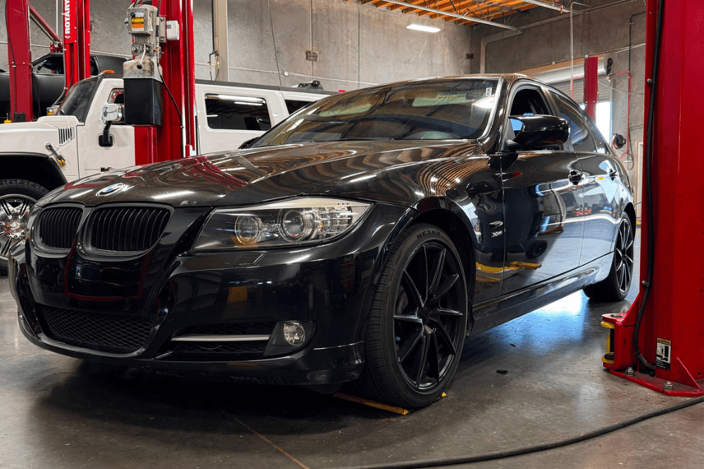 Black BMW 3 Series parked in APA Total Care Care's professional auto repair shop in Queen Creek, AZ, positioned between hydraulic lifts for inspection or European auto repair under high shop lighting.