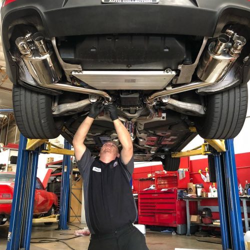 An auto repair technician working on the exhaust pipes of a dark grey vehicle